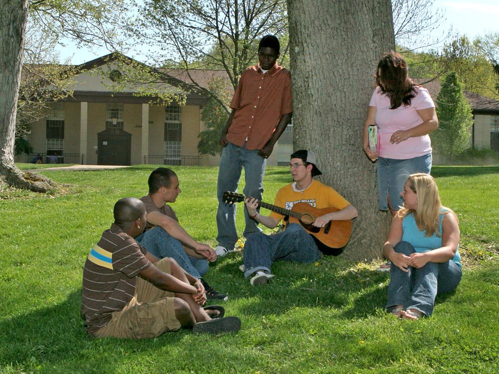 A group of six people sit and stand under a large tree. One person is playing a guitar, while others are listening and talking. A building is visible in the background.